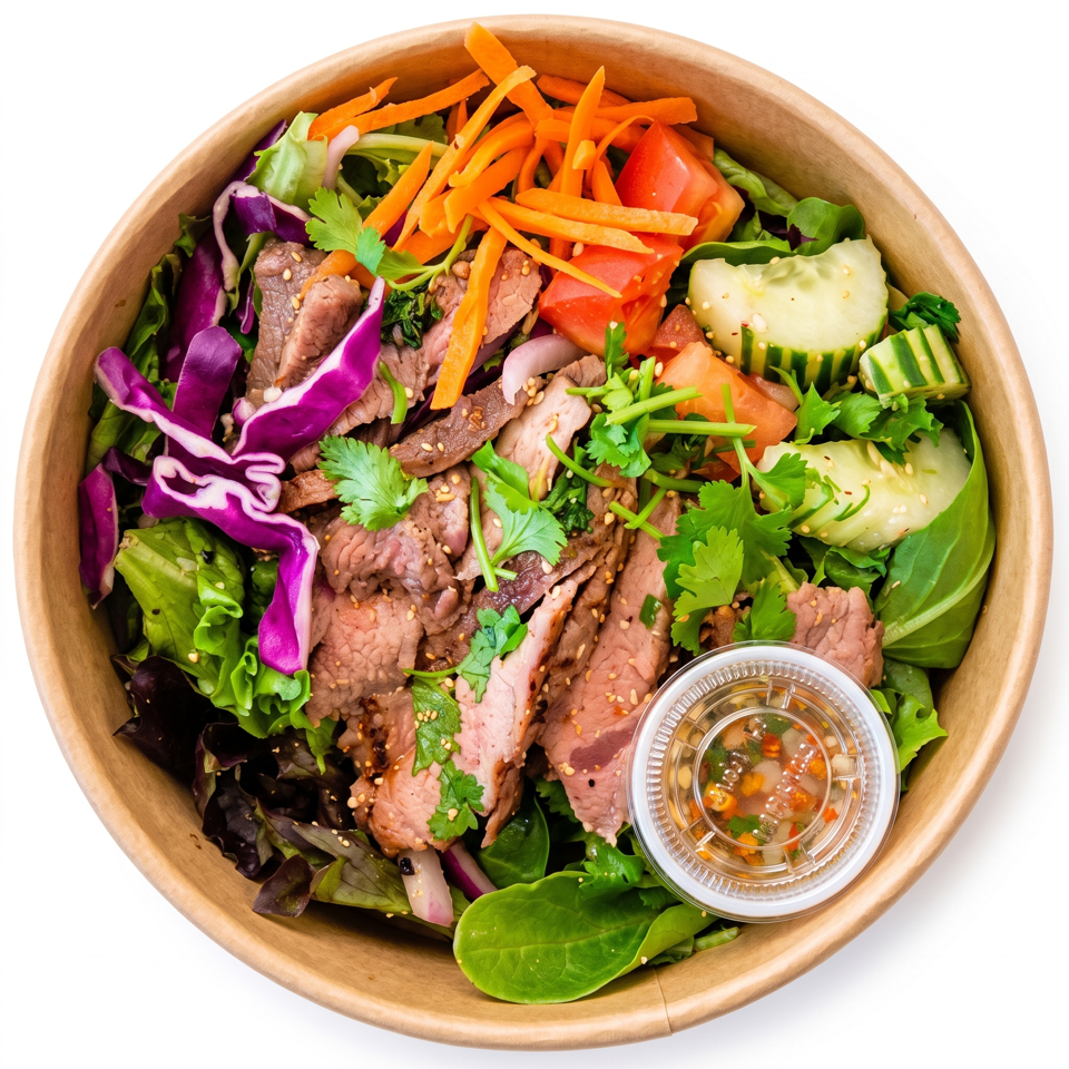 Colorful salad bowl with sliced beef, mixed greens, carrots, tomatoes, cucumber, cabbage, cilantro, sesame seeds, and dipping sauce.